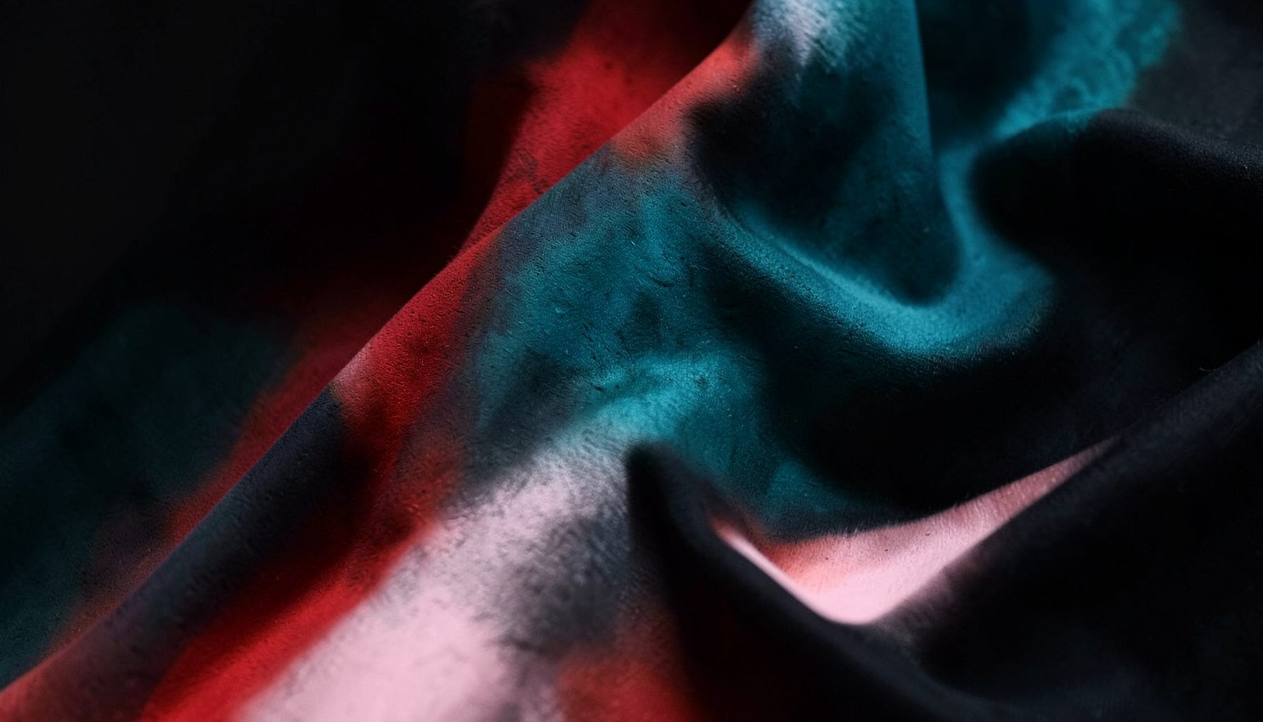 A close-up of richly textured hand-printed fabric in deep teal, black, and soft pink, draped in flowing folds against a dark background